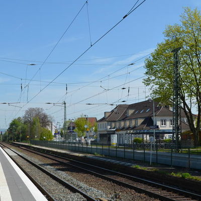 Das Foto wurde vom gegenüberliegenden Bahnsteig gemacht. Man sieht beide parallel verlaufenden Gleise im Hintergrund ist der Fahrradunterstand mit Glasdach, sowie das Bahnhofsgebäude zu sehen.