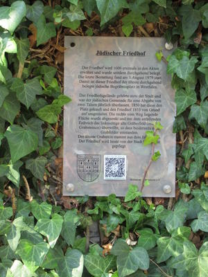 Das Bild zeigt eine Tafel mit Informationen zum jüdischen Friedhof, darauf neu angebracht ist ein QR-Code zum Scannen.